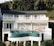 A modern white building features large windows and a curved balcony overlooking lush greenery. An infinity pool with teal tiles sits on an elevated platform. Several people are lounging on deck chairs and sitting by the poolside, enjoying the tranquil setting.