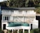 A modern white building features large windows and a curved balcony overlooking lush greenery. An infinity pool with teal tiles sits on an elevated platform. Several people are lounging on deck chairs and sitting by the poolside, enjoying the tranquil setting.