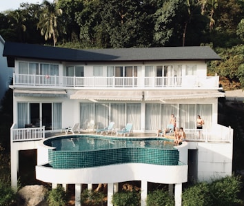 A modern white building features large windows and a curved balcony overlooking lush greenery. An infinity pool with teal tiles sits on an elevated platform. Several people are lounging on deck chairs and sitting by the poolside, enjoying the tranquil setting.