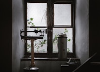 An old-fashioned balance scale sits on a windowsill of an aged wooden window with frosted glass panes. Ivy leaves are visible growing outside the window. There is a metal canister next to the scale, and dry foliage is hanging above the window.