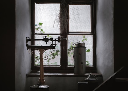 An old-fashioned balance scale sits on a windowsill of an aged wooden window with frosted glass panes. Ivy leaves are visible growing outside the window. There is a metal canister next to the scale, and dry foliage is hanging above the window.