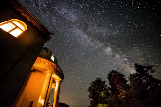 An observatory building with illuminated windows stands under a night sky filled with a dense array of stars and a visibly bright Milky Way. Tall trees silhouette against the sky, adding depth to the scene.