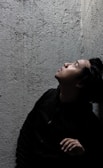A person in a dimly lit space looks upwards with a contemplative expression, surrounded by textured concrete walls.