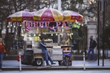 A street vendor is leaning against a colorful food cart adorned with umbrellas branded with Coca-Cola and NIKAS. The cart is lit with string lights and digital signs displaying neon text. Condiments and various food items are visible, along with small flags representing different countries. The scene includes a woman walking past and another person approaching the cart.
