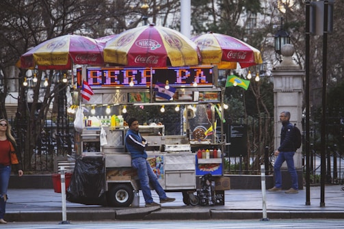 A street vendor is leaning against a colorful food cart adorned with umbrellas branded with Coca-Cola and NIKAS. The cart is lit with string lights and digital signs displaying neon text. Condiments and various food items are visible, along with small flags representing different countries. The scene includes a woman walking past and another person approaching the cart.