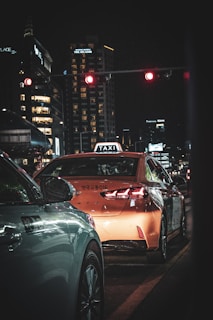 A nighttime urban scene with a distinctive focus on a taxi, identifiable by its rooftop sign, waiting at a red traffic light. The surrounding environment is characterized by tall city buildings illuminated with various lights, creating a dynamic backdrop. A car is also visible alongside the taxi, both stationary.