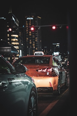 A nighttime urban scene with a distinctive focus on a taxi, identifiable by its rooftop sign, waiting at a red traffic light. The surrounding environment is characterized by tall city buildings illuminated with various lights, creating a dynamic backdrop. A car is also visible alongside the taxi, both stationary.