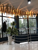 A modern, well-lit interior space featuring large windows with a view of trees outside. The ceiling has a unique light fixture composed of cylindrical wooden elements emitting soft, warm light. There is a coat stand on the left with a dark jacket hanging on it and a tall plant with broad leaves in a white pot. The floor has glossy white tiles, and a metallic railing adds a contemporary touch.