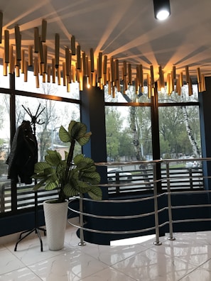 A modern, well-lit interior space featuring large windows with a view of trees outside. The ceiling has a unique light fixture composed of cylindrical wooden elements emitting soft, warm light. There is a coat stand on the left with a dark jacket hanging on it and a tall plant with broad leaves in a white pot. The floor has glossy white tiles, and a metallic railing adds a contemporary touch.