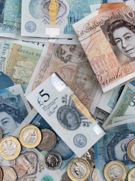 A collection of British banknotes and coins scattered in an overlapping arrangement. The banknotes include various denominations such as five and ten pounds, featuring iconic engravings and portrait art. Coins of varying sizes and colors accompany the notes, adding a metallic contrast to the paper currency.