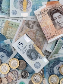 A collection of British banknotes and coins scattered in an overlapping arrangement. The banknotes include various denominations such as five and ten pounds, featuring iconic engravings and portrait art. Coins of varying sizes and colors accompany the notes, adding a metallic contrast to the paper currency.