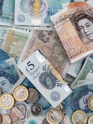 A collection of British banknotes and coins scattered in an overlapping arrangement. The banknotes include various denominations such as five and ten pounds, featuring iconic engravings and portrait art. Coins of varying sizes and colors accompany the notes, adding a metallic contrast to the paper currency.