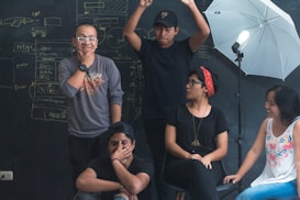 A group of five people are gathered in front of a chalkboard with sketches and notes written on it. One person is standing with arms raised, another is seated with a thoughtful expression, and others are engaged in conversation or posing playfully. A studio light with an umbrella is set up nearby.