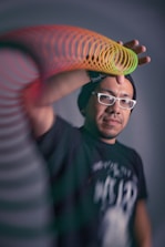 A person wearing a black beanie and white glasses is extending their hand with a colorful slinky toy. The background is blurred and the focus is on the subject's face and the slinky's vibrant colors.