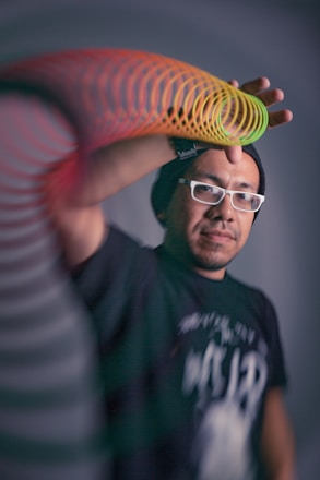 A person wearing a black beanie and white glasses is extending their hand with a colorful slinky toy. The background is blurred and the focus is on the subject's face and the slinky's vibrant colors.