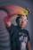 A person wearing a black beanie and white glasses is extending their hand with a colorful slinky toy. The background is blurred and the focus is on the subject's face and the slinky's vibrant colors.