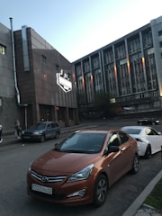 A street scene featuring parked cars with a prominent orange Hyundai in the foreground. Buildings with a modern architectural style are in the background, one has a large illuminated sign. The lighting suggests either early morning or late afternoon, with a clear sky overhead. A few pedestrians are visible on the sidewalk.