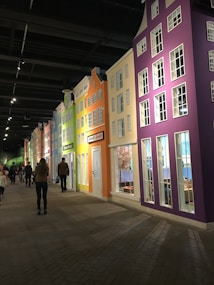 A brightly lit indoor display of colorful, narrow buildings styled like a quaint European street. Each building is painted in vibrant colors such as purple, orange, yellow, and green. There are several signs on the buildings indicating various businesses or attractions, including a flower market. The street is populated with a few people walking and observing the structures under a high ceiling with exposed beams and spotlighting.