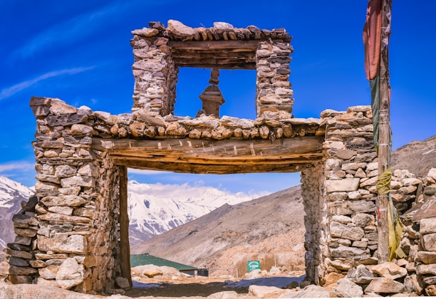 A stone structure stands against a backdrop of rugged, snow-capped mountains under a clear blue sky. The ancient-looking gateway is constructed from irregular stones and wooden beams. Nearby, a tall, colorful flag flutters on a pole.