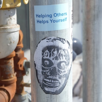 A metallic pole featuring a blue sticker with the phrase 'Helping Others Helps Yourself' and a black stencil art depiction of a face resembling a mask or skull.