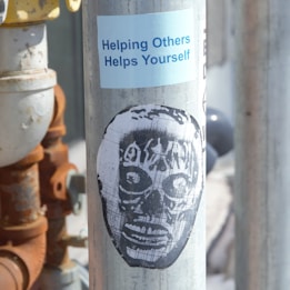 A metallic pole featuring a blue sticker with the phrase 'Helping Others Helps Yourself' and a black stencil art depiction of a face resembling a mask or skull.