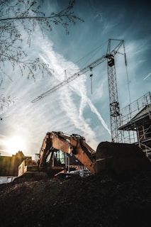 A construction site featuring a large excavator and a tall crane against a backdrop of a setting or rising sun. The sky is marked with dramatic cloud patterns and contrails. The equipment is silhouetted, with a nearby building partially visible. The scene includes bare branches in the foreground, adding depth and texture.