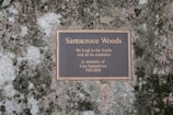 A memorial plaque mounted on a natural stone surface, inscribed with a dedication to Santacroce Woods, urging kindness to the Earth and its creatures.