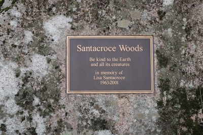 A memorial plaque mounted on a natural stone surface, inscribed with a dedication to Santacroce Woods, urging kindness to the Earth and its creatures.