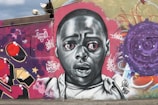 A vibrant graffiti mural featuring a realistic portrait of a wide-eyed, expressive person in grayscale. Surrounding the face are colorful abstract patterns and text, including graffiti tags and symbols. The art is set against a textured wall with shades of purple, red, and tan.