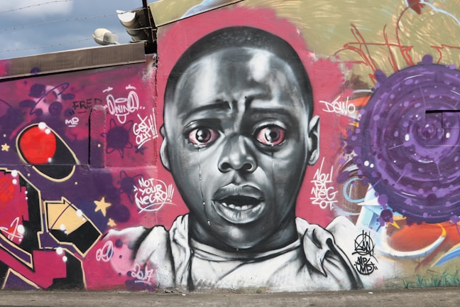 A vibrant graffiti mural featuring a realistic portrait of a wide-eyed, expressive person in grayscale. Surrounding the face are colorful abstract patterns and text, including graffiti tags and symbols. The art is set against a textured wall with shades of purple, red, and tan.