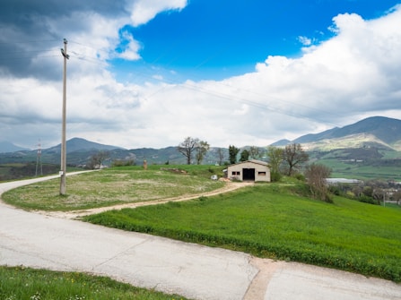 A small rural building sits on a grassy hill surrounded by fields and distant mountains under a partially cloudy sky. Power lines run across the scene, and a winding paved road leads toward the building.