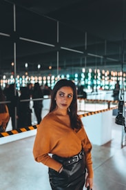 A woman stands confidently in front of a mirrored wall reflecting numerous small lights. She is wearing a stylish orange top and black leather skirt with a belt. In the background, the lights create a dazzling pattern enhanced by the reflective surfaces, contributing to a modern and sophisticated atmosphere.