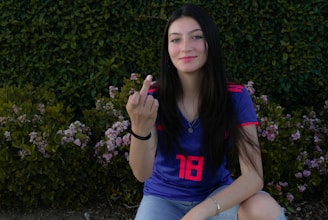 A person with long dark hair is sitting in front of a lush green hedge with small pink flowers. They are wearing a blue sports jersey with red accents, displaying the number 18. The person has a slight smile and is holding up one hand with the middle finger extended.
