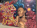 A colorful mural featuring a portrait of a woman with her eyes closed, surrounded by roses and cosmic elements. The mural includes bold, flowing script that reads 'Believe in the Reality of Your Dreams' in vibrant colors.