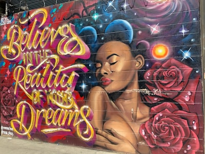 A colorful mural featuring a portrait of a woman with her eyes closed, surrounded by roses and cosmic elements. The mural includes bold, flowing script that reads 'Believe in the Reality of Your Dreams' in vibrant colors.