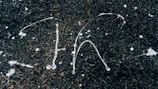White paint splatters on a rough, dark asphalt surface creating abstract patterns. The contrast between the bright paint and the dark background highlights the textured surface.