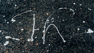 White paint splatters on a rough, dark asphalt surface creating abstract patterns. The contrast between the bright paint and the dark background highlights the textured surface.
