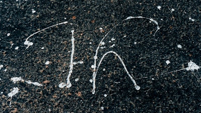 White paint splatters on a rough, dark asphalt surface creating abstract patterns. The contrast between the bright paint and the dark background highlights the textured surface.