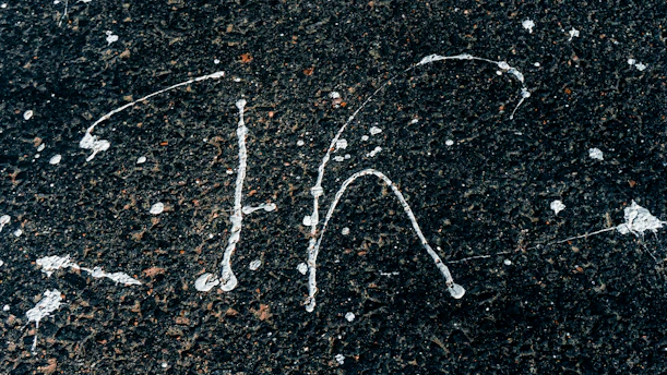White paint splatters on a rough, dark asphalt surface creating abstract patterns. The contrast between the bright paint and the dark background highlights the textured surface.