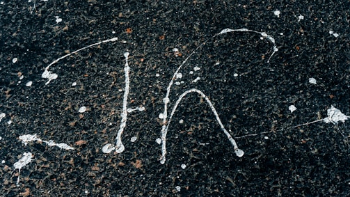 White paint splatters on a rough, dark asphalt surface creating abstract patterns. The contrast between the bright paint and the dark background highlights the textured surface.