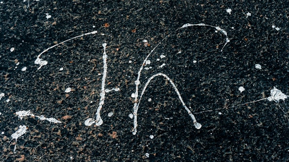 White paint splatters on a rough, dark asphalt surface creating abstract patterns. The contrast between the bright paint and the dark background highlights the textured surface.