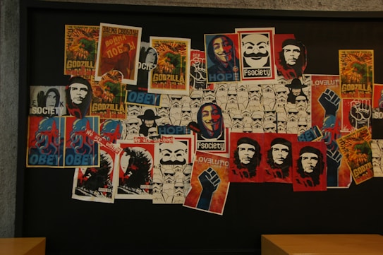 A collage of posters and images on a wall, featuring a mix of pop culture and political references. Notable elements include depictions of Godzilla, the 'Obey' motif with an octopus, Che Guevara's portrait, and the masked, smiling face associated with 'F Society' and the popular anonymous mask. Stormtrooper graphics are present, as well as various posters with slogans and symbols of resistance.