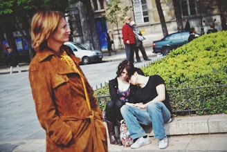 A woman in a brown coat walks by in the foreground while, in the background, a young couple sits closely together on a stone bench beside a green shrub, with the man resting his head on the woman's shoulder. The setting is an outdoor urban area with people and parked cars visible in the street along with trees and a historic building.