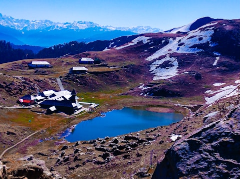 A serene mountainous landscape with a tranquil lake surrounded by rocky terrain. Snow-dappled hills stretch into the distance, with several quaint buildings scattered around the lake. In the background, majestic, snow-capped mountains rise under a clear blue sky.