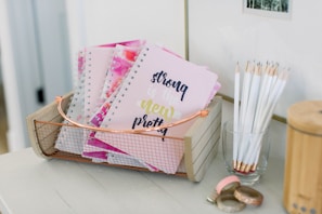 A wooden basket holds multiple spiral notebooks with pastel pink covers, featuring the text 'strong is the new pretty' in decorative fonts. Near the basket, a transparent glass is filled with white pencils, all sharpened and standing upright. On the table, there are decorative rolls of washi tape with metallic and glittery designs.