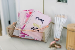 A wooden basket holds multiple spiral notebooks with pastel pink covers, featuring the text 'strong is the new pretty' in decorative fonts. Near the basket, a transparent glass is filled with white pencils, all sharpened and standing upright. On the table, there are decorative rolls of washi tape with metallic and glittery designs.