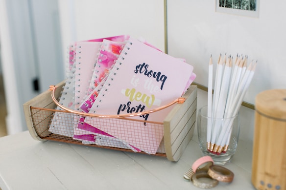 A wooden basket holds multiple spiral notebooks with pastel pink covers, featuring the text 'strong is the new pretty' in decorative fonts. Near the basket, a transparent glass is filled with white pencils, all sharpened and standing upright. On the table, there are decorative rolls of washi tape with metallic and glittery designs.