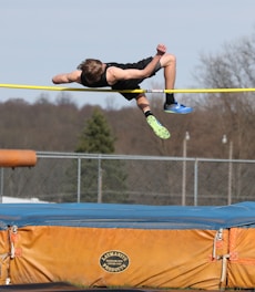 An athlete is performing a high jump over a horizontal bar. The athlete is in mid-air above a blue and orange landing mat. The background features a fenced area with trees and a clear sky.