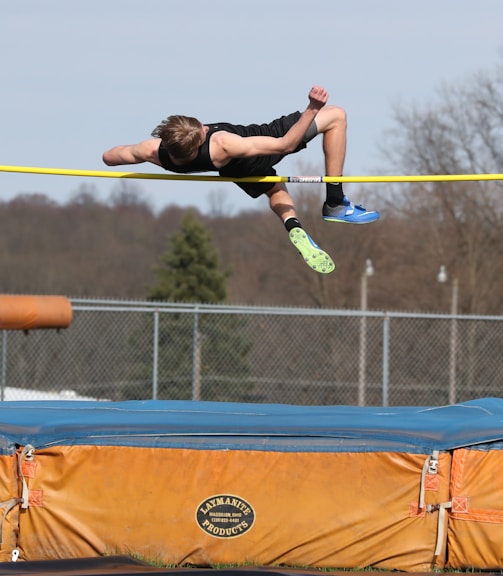 An athlete is performing a high jump over a horizontal bar. The athlete is in mid-air above a blue and orange landing mat. The background features a fenced area with trees and a clear sky.