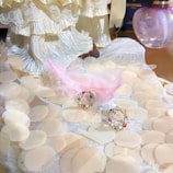 A delicate arrangement featuring a pink feather, lace fabric, and a pair of circular earrings adorned with gemstones, set on a surface with translucent, round embellishments. A bottle of perfume sits in the background with decorative attire partially visible.